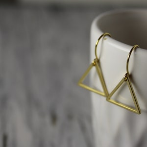 Könnte beinhalten: Ein Paar goldene Dreiecksohrringe mit schlichtem Design. Die Ohrringe hängen an einer weißen Keramiktasse.