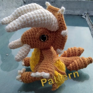 May include: A crocheted amigurumi toy in the shape of a brown and yellow dinosaur with white horns and a black eye. The toy is sitting on a blue and white patterned surface. The word "Pattern" is written in yellow on the surface.