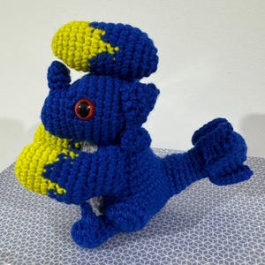 Könnte beinhalten: Blau-gelbes Häkeltier in Form eines Drachen. Der Drache hat einen großen Kopf mit einem einzigen roten Auge und einen langen Schwanz.