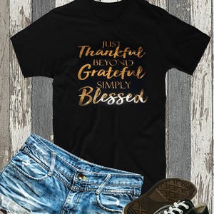 以下が含まれることがあります： 黒いTシャツに、金色の箔押しで「Just Thankful Beyond Grateful Simply Blessed」と書かれています。Tシャツはデニムのショートパンツと黒いスニーカーと合わせています。