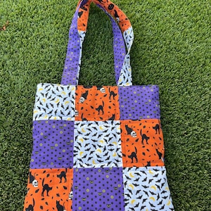 May include: A patchwork tote bag with a Halloween theme. The bag features a variety of patterned squares in orange, purple, and white. The patterns include black cats, bats, skulls, and polka dots. The handles are purple and orange.