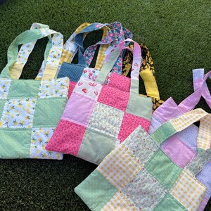 May include: A collection of handmade tote bags with patchwork designs. The bags feature various patterns, including lemons, strawberries, floral prints, and gingham. The color palette includes green, pink, yellow, and blue, with matching fabric handles.