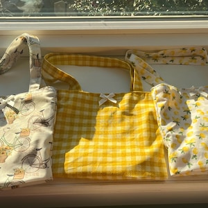 Może przedstawiać: Trzy torby typu tote z różnymi wzorami. Pierwsza torba jest biała z kwiatowym wzorem i rowerami. Druga torba jest w kratę żółto-białą. Trzecia torba jest biała z wzorem cytrynowym.
