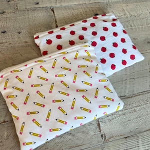 Op de afbeelding: Twee witte etuis met ritssluiting. Eén etui heeft een patroon van rode appels, de andere heeft een patroon van gele potloden met roze gummen. Beide etuis liggen op een licht houten oppervlak.