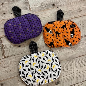 May include: Three quilted fabric pumpkin-shaped pot holders with black and white polka dot stems. One is purple with yellow cat eyes, one is orange with black cats and skulls, and one is white with black bats and yellow crescent moons.