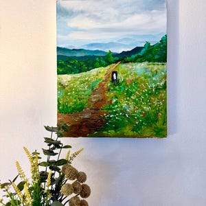 May include: An oil painting depicts a landscape with a path winding through a field of white flowers. The path leads to a distant mountain range under a cloudy sky. The artwork is framed and mounted on a white wall.