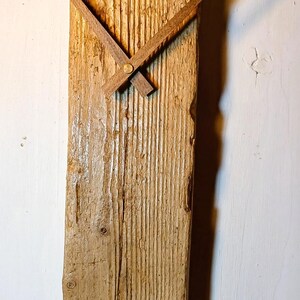 May include: A vertical wall clock crafted from a single, weathered piece of light brown wood with a prominent grain and natural texture. The clock features two simple, straight wooden hands attached to a central brass-coloured pin.