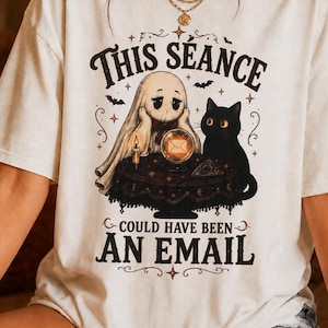 Könnte beinhalten: Ein cremefarbenes T-Shirt mit kurzen Ärmeln und einer Grafik eines Geistes und einer schwarzen Katze, die an einem Tisch mit einer Kristallkugel sitzen. Der Text lautet THIS SEANCE COULD HAVE BEEN AN EMAIL. Das Design zeigt kleine Fledermäuse.