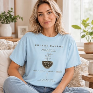 Könnte beinhalten: Ein hellblaues T-Shirt mit dunklem Text und einer Grafik einer Pflanze, die aus einer gesprungenen Schale wächst. Der Text lautet: EHLERS DANLOS, NOT A FLAW IN ME, A DIFFERENT DESIGN, fragile + resilient + adaptable + unbreakable, IT'S NOT IN MY HEAD, IT'S IN MY CONNECTIVE TISSUE.