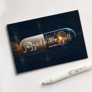 Pode incluir: Uma impressão retangular com uma cápsula de lado esquerdo cinzento texturizado e lado direito azul translúcido contendo padrões neuronais dourados brilhantes. O texto "jagged little pill" está escrito em branco. Uma caneta de gel branca.