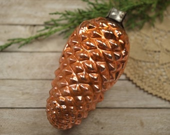 May include: A copper-colored glass Christmas ornament shaped like a pine cone. The ornament has a shiny, metallic finish and is detailed with a textured surface.