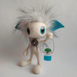 Peut inclure: Une créature en feutre fantaisiste avec une tête blanche et grise duveteuse, de grands yeux bleus et des accents turquoise. Elle tient une petite bouteille en verre avec un bouchon vert et un liquide bleu. La créature a un bouton argenté sur la poitrine.