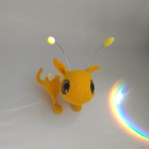 Può includere: Una creatura fatta a mano in feltro giallo con grandi occhi espressivi e piccole zampe. Ha due antenne, una con un globo giallo luminoso. La creatura ha accenti bianchi sulla schiena e un design stravagante e da cartone animato.