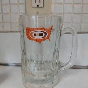 May include: Clear glass beer mug with a handle. The mug features a rectangular design and an orange and brown sticker with the A&W logo and a map of the United States.