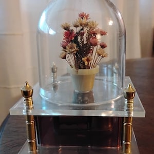 May include: A decorative floral arrangement encased in a clear glass dome. The flowers, in shades of pink, yellow, and brown, are in a small, light yellow pot. The dome sits on a clear acrylic base with gold-colored accents.