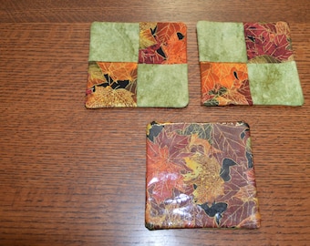 Puede incluir: Tres posavasos cuadrados de tela con un patrón de hojas de otoño. Dos posavasos son verdes con un mosaico de hojas de otoño en naranja, rojo y amarillo. El tercer posavasos es un patrón sólido de hojas de otoño en naranja, rojo y amarillo.