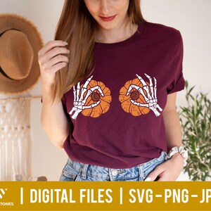 May include: A maroon t-shirt with a graphic of two skeleton hands holding pumpkins. The pumpkins are orange with a brown stem and a black seed inside.