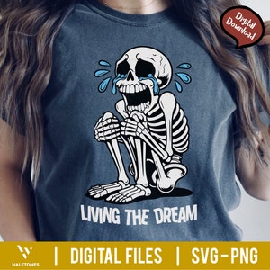 May include: A dark blue t-shirt featuring a crying skeleton illustration. The skeleton is white with black outlines, and blue tears are streaming from its eyes. The text "LIVING THE DREAM" is printed in white below the skeleton.