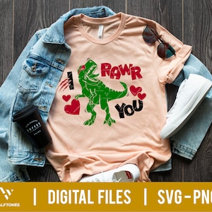 May include: A light pink t-shirt with a green dinosaur graphic and the text "I Rawr You". The dinosaur is wearing a red headband and has hearts around it.