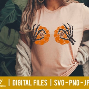 May include: A pink t-shirt with a graphic of two orange pumpkins with black skeletal hands holding them up to their eyes. The pumpkins have black eyes and a black stem.