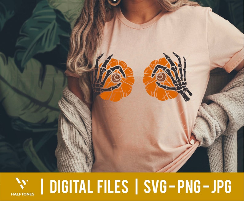 May include: A pink t-shirt with a graphic of two orange pumpkins with black skeletal hands holding them up to their eyes. The pumpkins have black eyes and a black stem.