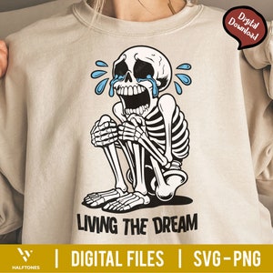 May include: Beige sweatshirt featuring a crying skeleton illustration with blue tears. The skeleton is in a seated position with its hands clasped. The text "LIVING THE DREAM" is below the illustration. The words "DIGITAL FILES" and "SVG-PNG" are also present.