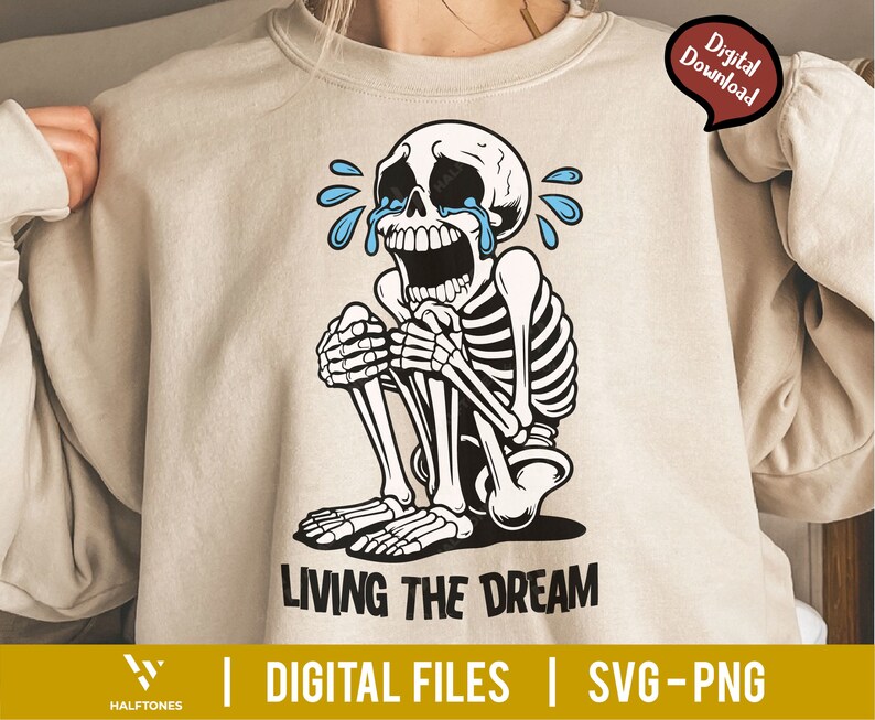 May include: Beige sweatshirt featuring a crying skeleton illustration with blue tears. The skeleton is in a seated position with its hands clasped. The text "LIVING THE DREAM" is below the illustration. The words "DIGITAL FILES" and "SVG-PNG" are also present.