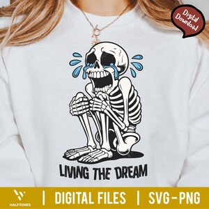 May include: White sweatshirt featuring a crying skeleton illustration with blue tears. The skeleton is sitting with its knees up, and the text "LIVING THE DREAM" is below. The image also includes the text "DIGITAL FILES | SVG-PNG".