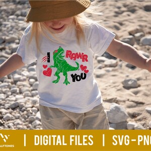 May include: A white t-shirt with a green dinosaur graphic and the text "I Roar You" in red and black letters. The dinosaur is holding a red heart in its mouth.
