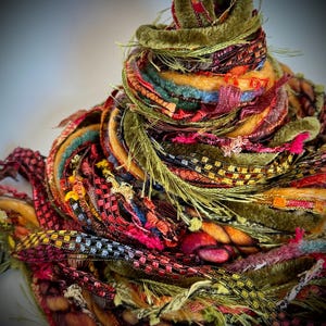 May include: A conical bundle of assorted decorative ribbons and yarns in shades of olive green, deep red, orange, and gold. The collection features various textures including velvet, metallic woven strips, frayed edges, and twisted wool fibers.