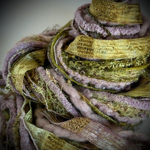 May include: Close-up of a textured art piece featuring a mix of materials. The piece incorporates shades of lavender, olive green, and gold. The design includes ribbons, yarn, and woven elements, creating a layered and tactile appearance.