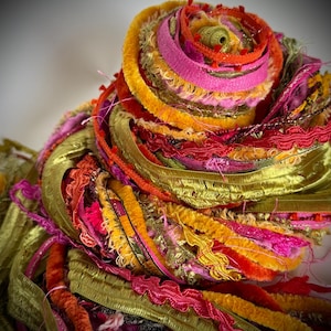 May include: A coiled bundle of assorted textile ribbons and trims in vibrant shades of olive green, hot pink, bright orange, and deep red. The collection features various textures including velvet, metallic, ruffled, and frayed fabric strips.