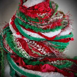 May include: A decorative bundle of assorted yarn and ribbon in red, green, and white. The collection features various textures including fuzzy white chenille, smooth red satin, green velvet, and metallic silver threads woven throughout the mix.