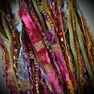May include: A collection of assorted decorative ribbons and yarn strands in various textures and colours, including shades of magenta, olive green, gold, and light blue. The bundle features velvet, metallic, and ruffled trims layered together.
