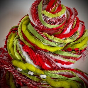 May include: A decorative bundle of assorted yarn and ribbon in shades of lime green, bright red, and white. The collection features various textures including fuzzy wool, metallic silver threads, smooth satin ribbons, and frayed fabric strips.