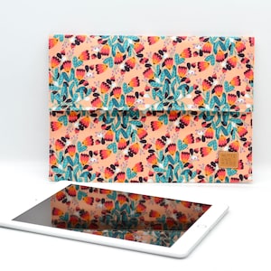 May include: A pink floral tablet sleeve with a brown leather tag that says "Made with Love in NYC". The sleeve is lying on a white surface next to a white tablet.