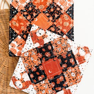 May include: Two quilted Halloween-themed pot holders. The pot holders feature a patchwork design with orange pumpkins, black floral patterns, and white accents. One pot holder has a square shape, and the other is diamond-shaped.