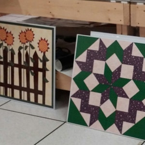 May include: Two framed quilt squares. One square features a brown picket fence with three orange sunflowers. The other square features a green and purple starburst pattern.