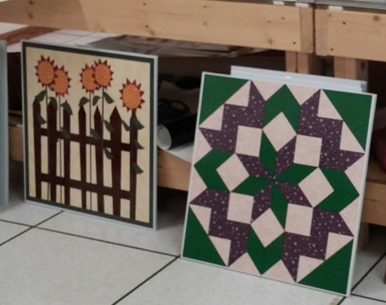 May include: Two framed quilt squares. One square features a brown picket fence with three orange sunflowers. The other square features a green and purple starburst pattern.