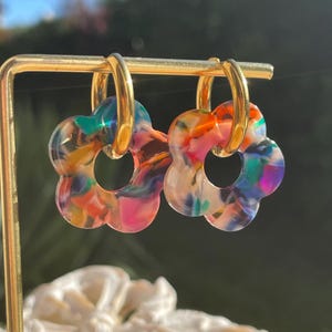 Op de afbeelding: Een paar bloemvormige oorbellen met gouden ringen. De oorbellen hebben een kleurrijk, gemarmerd ontwerp met tinten roze, oranje, blauw en groen. De oorbellen hangen aan een gouden displaystandaard.