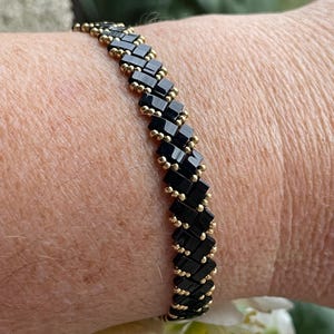 Op de afbeelding: Een zwarte en gouden kralen armband met een chevron patroon. De armband wordt gedragen op een pols.