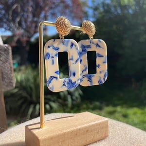 May include: A pair of statement earrings with a cream-colored rectangular design, featuring blue brushstroke patterns. The earrings have gold-colored stud tops and are displayed on a gold stand with a wooden base. The background is blurred with greenery.