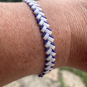 Peut inclure: Un bracelet de perles blanc et bleu. Le bracelet présente un motif répétitif de perles rectangulaires blanches séparées par de petites perles bleues. Le bracelet est porté au poignet.