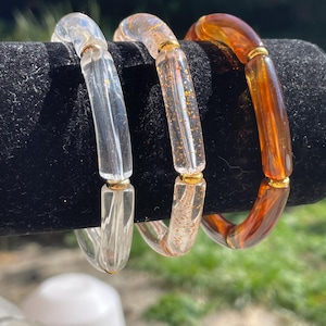 Peut inclure: Trois bracelets rigides en plastique transparent avec des accents dorés. Les bracelets sont empilés sur une surface noire. Le bracelet du milieu a des paillettes dorées incrustées dans le plastique.