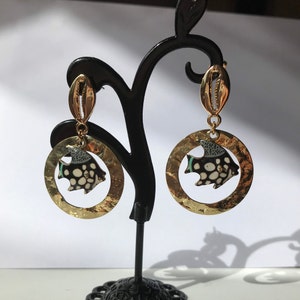 May include: Gold dangle earrings with a black and white fish design. The fish has a green and white pattern on its body. The earrings are hanging from a gold hook.