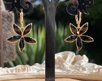 May include: A pair of black flower-shaped earrings with gold-colored trim. The earrings are displayed on a black earring stand. The earrings have a hoop closure. The background is blurred, with hints of greenery and sunlight.