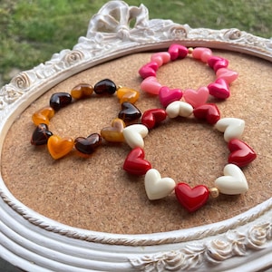 Peut inclure: Trois bracelets de perles en forme de cœur, dans des tons de marron, rose, rouge et blanc. Les bracelets sont disposés sur un tableau en liège, encadré d'un cadre blanc décoratif. Chaque bracelet est composé de perles en forme de cœur.