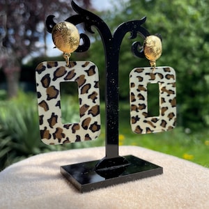 Peut inclure: Une paire de boucles d'oreilles rectangulaires en imprimé léopard marron et or avec des accents ronds dorés.