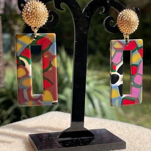 Op de afbeelding: Een paar kleurrijke rechthoekige oorbellen met een gouden, getextureerde bal aan de bovenkant. De oorbellen worden getoond op een zwarte standaard.