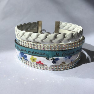 Puede incluir: Un brazalete de cuero trenzado blanco con una banda turquesa que dice "Je t'aime maman" y una banda blanca con pedrería. El brazalete también tiene una banda de tela blanca con un estampado floral y un dije de estrella plateada.